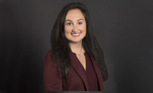 Portrait-style headshot of woman with long dark brown hair, wearing a maroon outfit and sitting against a dark brown background.
